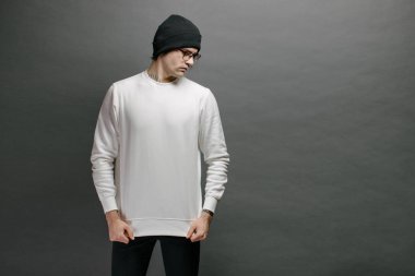 Beyaz kazak giyen ve gri arka planda duran siyah şapkalı bir adam. Tişört ya da kapüşonlu süveter. Model, logo tasarımları ya da boş alanı olan tasarım baskıları..