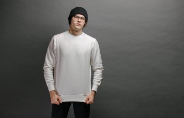 Beyaz kazak giyen ve gri arka planda duran siyah şapkalı bir adam. Tişört ya da kapüşonlu süveter. Model, logo tasarımları ya da boş alanı olan tasarım baskıları..