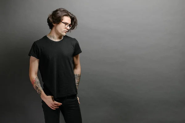 Hipster handsome male model with glasses wearing black blank t-shirt and black jeans with space for your logo or design in casual urban style — Stock Photo, Image