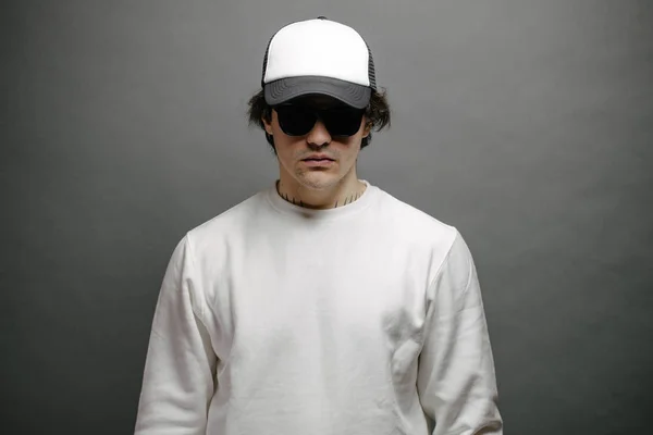 Man wearing blank white sweatshirt and empty baseball cap standing over gray background. Sweatshirt or hoodie for mock up, logo designs or design print with with free space.