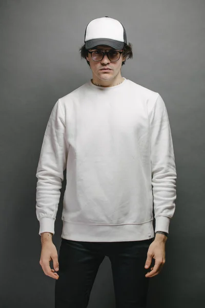 Man wearing blank white sweatshirt and empty baseball cap standing over gray background. Sweatshirt or hoodie for mock up, logo designs or design print with with free space.