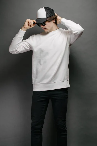 Man wearing blank white sweatshirt and empty baseball cap standing over gray background. Sweatshirt or hoodie for mock up, logo designs or design print with with free space.