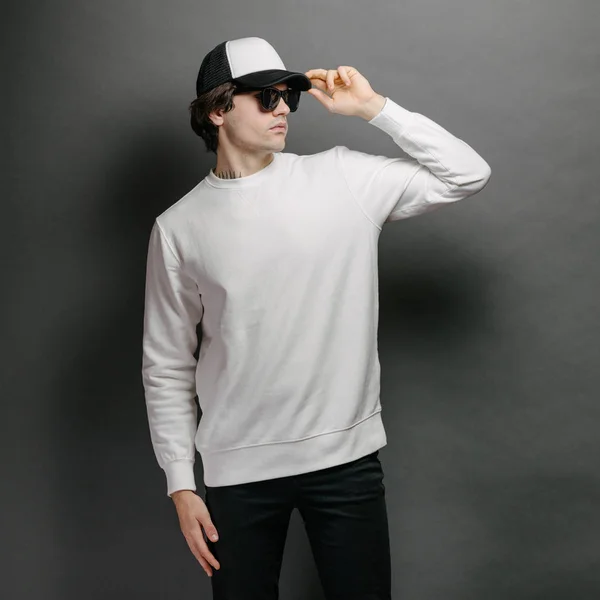 Homem vestindo camisola branca em branco e boné de beisebol vazio em pé sobre fundo cinza. Camisola ou capuz para simular, desenhos de logotipo ou impressão de design com espaço livre. — Fotografia de Stock