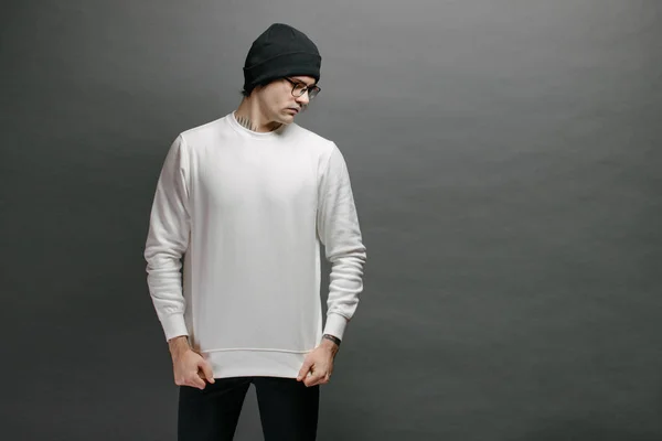 Een man met een witte sweater en een zwarte hoed over een grijze achtergrond. Sweatshirt of hoodie voor mock up, logo designs of design prints met vrije ruimte. — Stockfoto