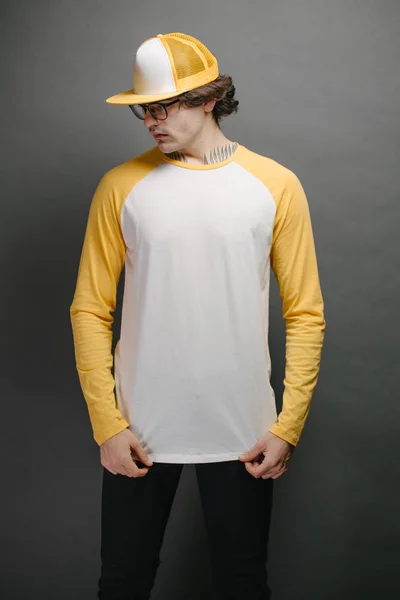 흰색과 노란색이 섞인 롱 슬리브 셔츠를 입은 남자 모델, 회색 배경 위에 로고나 디자인을 위한 공간이 있는 야구 모자를 착용 한 남자 모델 — 스톡 사진
