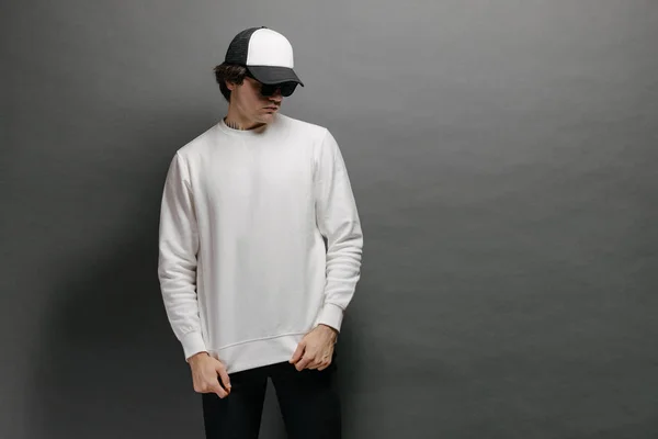 Hombre con sudadera blanca en blanco y gorra de béisbol vacía de pie sobre fondo gris. Sudadera o sudadera con capucha para maqueta, diseños de logotipo o impresión de diseño con espacio libre. —  Fotos de Stock