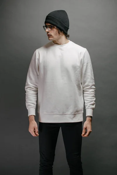 Een man met een witte sweater en een zwarte hoed over een grijze achtergrond. Sweatshirt of hoodie voor mock up, logo designs of design prints met vrije ruimte. — Stockfoto