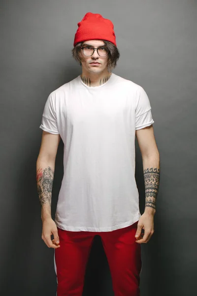 Hipster guapo modelo masculino con gafas con blanco blanco camiseta en blanco y pantalones rojos y un sombrero rojo con espacio para su logotipo o diseño sobre fondo gris —  Fotos de Stock