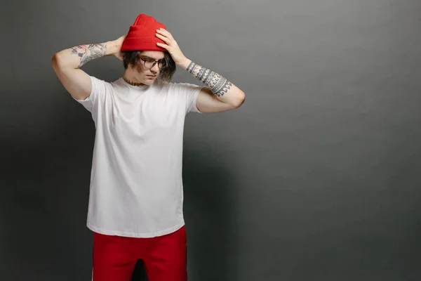 Hipster guapo modelo masculino con gafas con blanco blanco camiseta en blanco y pantalones rojos y un sombrero rojo con espacio para su logotipo o diseño sobre fondo gris —  Fotos de Stock