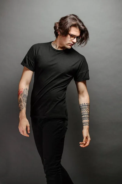 Hipster handsome male model with glasses wearing black blank t-shirt and black jeans with space for your logo or design in casual urban style — Stock Photo, Image