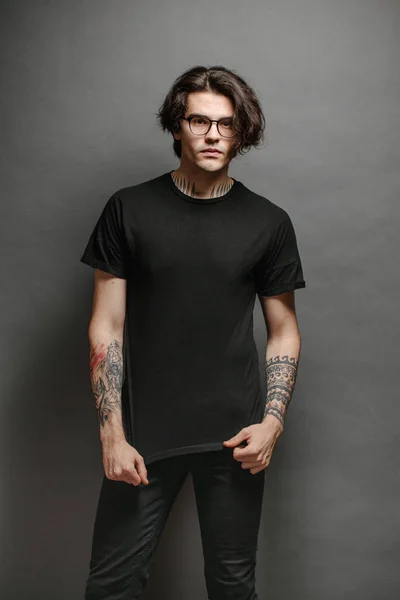 Hipster handsome male model with glasses wearing black blank t-shirt and black jeans with space for your logo or design in casual urban style — Stock Photo, Image