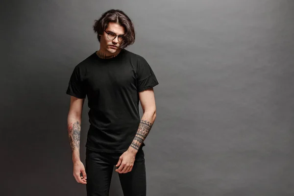 Hipster handsome male model with glasses wearing black blank t-shirt and black jeans with space for your logo or design in casual urban style — Stock Photo, Image