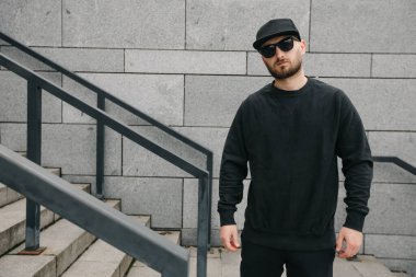 Beyaz kazak giyen ve beton duvarın üzerinde duran siyah şapkalı bir adam. Tişört ya da kapüşonlu süveter. Model, logo tasarımları ya da boş alanı olan tasarım baskıları.