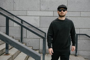 Beyaz kazak giyen ve beton duvarın üzerinde duran siyah şapkalı bir adam. Tişört ya da kapüşonlu süveter. Model, logo tasarımları ya da boş alanı olan tasarım baskıları.