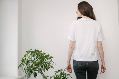Beyaz boş tişört giyen sıska bir kadın ve logonuz için yeri olan siyah kot pantolon giyen ya da beyaz duvarın üzerinde gelişigüzel şehir tarzı bir tasarım yapan bir kadın.