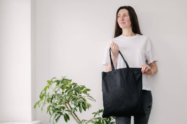 Beyaz duvara siyah tekstil eko çanta tutan genç bir kadın. Ekoloji ya da çevre koruma kavramı. Model için beyaz eko çanta.