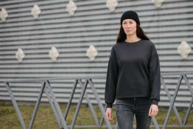Siyah eşofman giyen bir kadın gri arka planda, şehir merkezinde sıradan bir şekilde dikiliyor. Tişört ya da kapüşonlu süveter. Model, logo tasarımları ya da boş alanı olan tasarım baskıları.