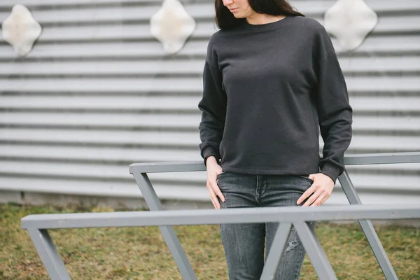 Woman wearing black sweatshirt standing in urban area in casual urban style over gray background. Sweatshirt or hoodie for mock up, logo designs or design prints with with free space