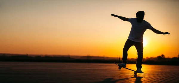 帽子付きのジーンズと白いTシャツを着て日没の間に街の通りのボードに乗っているスケーターのヒップスター男性 テキストの空のスペース — ストック写真