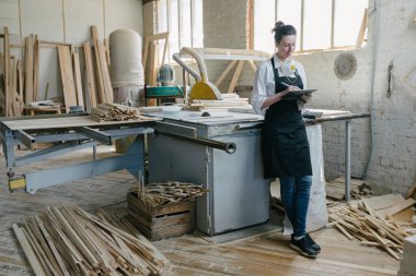 Kendi marangozhanesinde marangoz olarak çalışan kendine güvenen bir kadın. Bir tablet bilgisayar kullanıyor ve çalışma alanındayken notlar yazıyor. Küçük işletme kavramı.