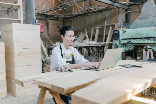 自信的女人在自己的木材店里做木匠 她在工作时使用笔记本电脑并写笔记 小企业概念 — 图库照片