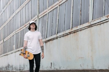 Sakallı, beyaz tişörtlü yakışıklı bir erkek manken ve logonuz için geniş bir beyzbol şapkası ya da gelişigüzel şehir tarzı bir tasarım. Elinde bir kaykay tutuyor.