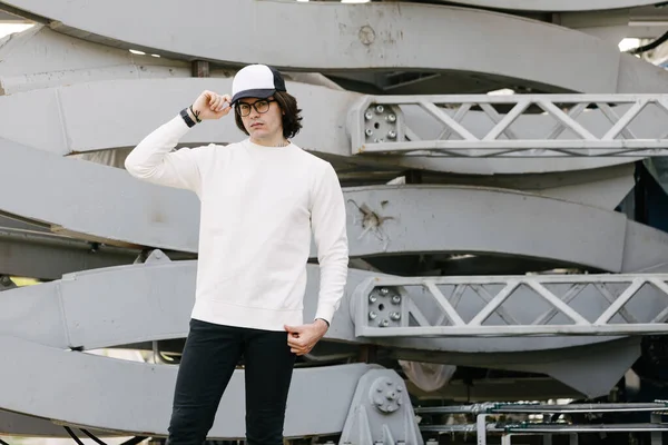 Man wearing blank white sweatshirt and empty baseball cap standing over gray background. Sweatshirt or hoodie for mock up, logo designs or design print with free space.