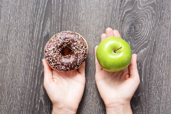 Gezonde levensstijl met groene appel en smakelijke donut in vrouw handen op houten achtergrond — Stockfoto