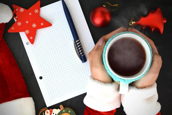 Vrouw in Santas kleren holding een kop hete thee over Kerstmis achtergrond, in de buurt van een lege lijst van de wens — Stockfoto