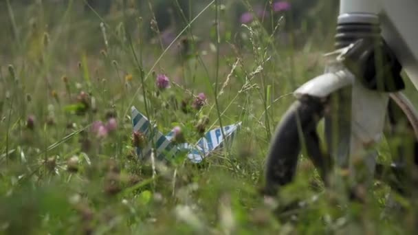 Hand van kind neemt papieren boot in groen gras in de buurt van fietswiel in het veld. — Stockvideo