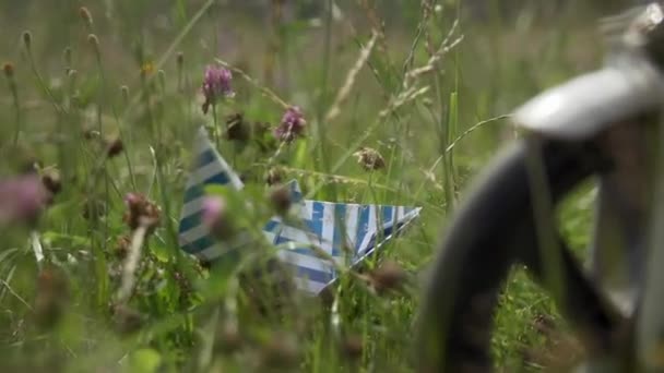 Hand of child takes paper boat in green grass near bicycle wheel in field. — Stock video