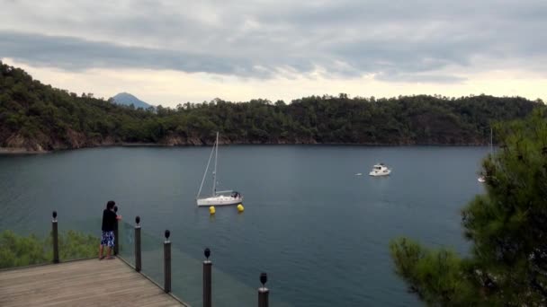 Pojken står på bron och tittar på båtar och berg på kusten i Bay. — Stockvideo