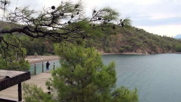Jongen staat op de brug op de achtergrond van de zee en bergen op de kust in de baai. — Stockvideo