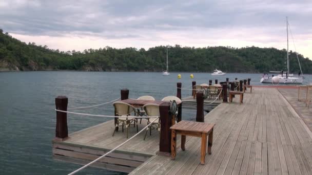 Stoly a židle na dřevěném molu na pozadí horského pobřeží moře. — Stock video
