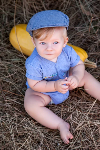 Little boy sits near a haystack in a blue dress and hat, lying next kabachek — Stock Photo, Image