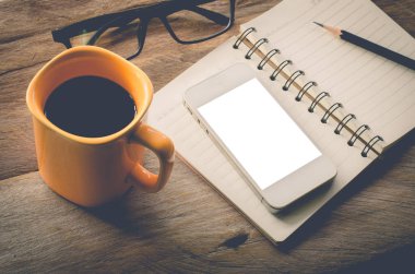 Kitaplı sarı kahve fincanı. Gözlük, not defteri. Ahşap masada telefon ve kalem.