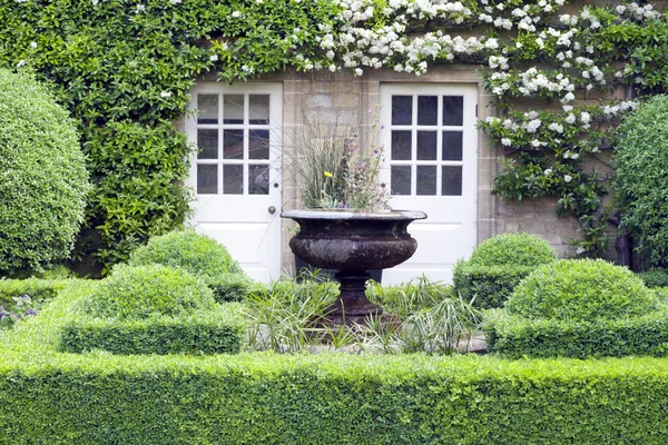 Topiary garden with ornamental plant pot in the middle, in front of stone cottage . — Stock Photo, Image