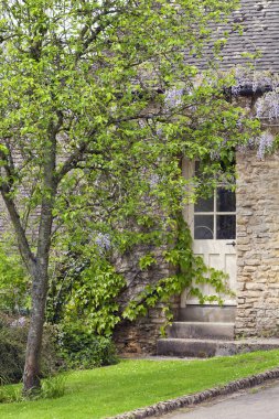 Kapılar bir eski, geleneksel Cotswolds taş yazlık mor çiçekli wisteria altında .