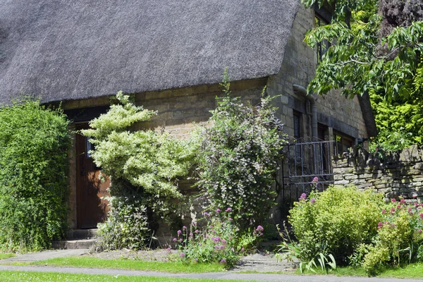 Secluded old stone cottage with thatched roof, iron gate on a side among flowers, climbing trees and shrubs in the front .