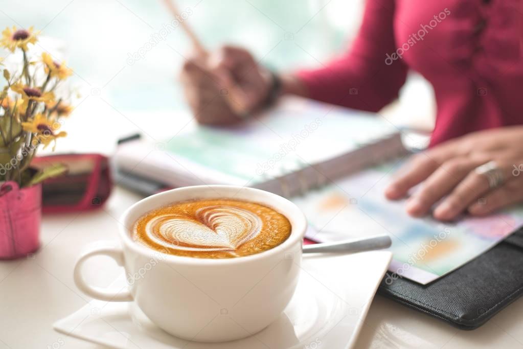 Coffee with Heart Shape Latte Art on Working Desk. Moody Shot, blur working woman checking figure in her chart in books. Concept passion work.