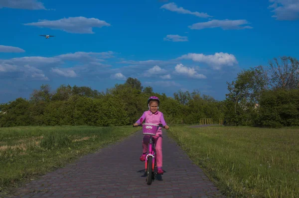 Fille dans un casque rose en vélo dans le parc, en arrière-plan dans le ciel un avion vole — Photo