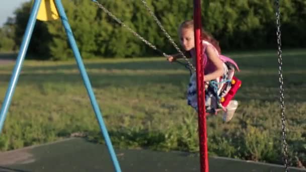 Menina alegre de sete anos em um vestido rosa balançando em um balanço no parque ao pôr do sol — Vídeo de Stock