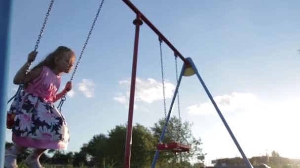 Menina alegre de sete anos em um vestido rosa balançando em um balanço no parque ao pôr do sol — Vídeo de Stock