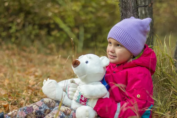 Une petite fille de sept ans assise sur un banc dans un parc d'automne, tenant son jouet préféré sur ses mains - un ours en peluche — Photo