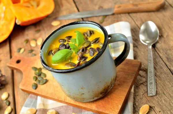 Starý smaltovaný šálek smetanové dýňové polévky s čerstvým bazalkou, dýňovými semínky, lžící a nožem na venkovském dřevěném stole — Stock fotografie