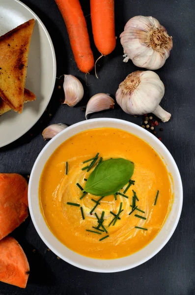 Schüssel Süßkartoffelcremesuppe mit frischem Basilikum und Schnittlauch, farbigem Pfeffer, Karotten, Knoblauch und geröstetem Brot auf Schieferplatte. Ansicht von oben. vertikales Foto. — Stockfoto