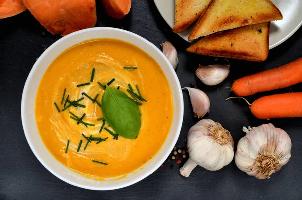 Чаша сладкого картофельного сливочного супа со свежим базиликом, а также перец, морковь и чеснок на сланцевой плите. Вид сверху . — стоковое фото