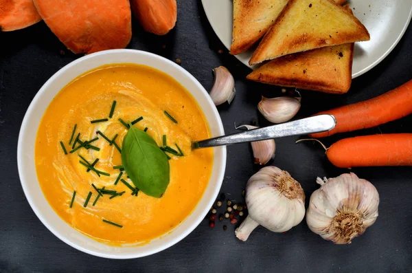 Schüssel Süßkartoffelcremesuppe mit frischem Basilikum, Schnittlauch und Löffel, farbigem Pfeffer, Karotten und Knoblauch auf Schieferplatte. Ansicht von oben. — Stockfoto