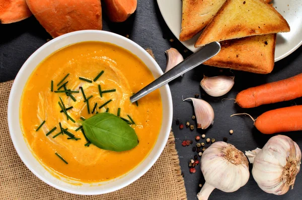 Schüssel Süßkartoffelcremesuppe mit frischem Schnittlauch und Basilikum und Löffel, Pfeffer, Karotte, Knoblauch und geröstetem Toastbrot auf Schieferplatte. Ansicht von oben. — Stockfoto