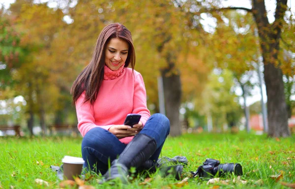 Modieuze tiener meisje met smartphone, camera en afhaalmaaltijden koffie in park in het najaar zit aan het glimlachen. Vrouw in herfst in park texting — Stockfoto